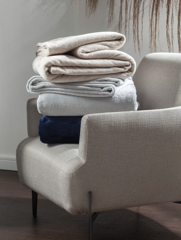 Pilha de cobertores Kacyumara Blanket 300 dobrados sobre uma poltrona, em tons de bege, cinza claro e azul marinho