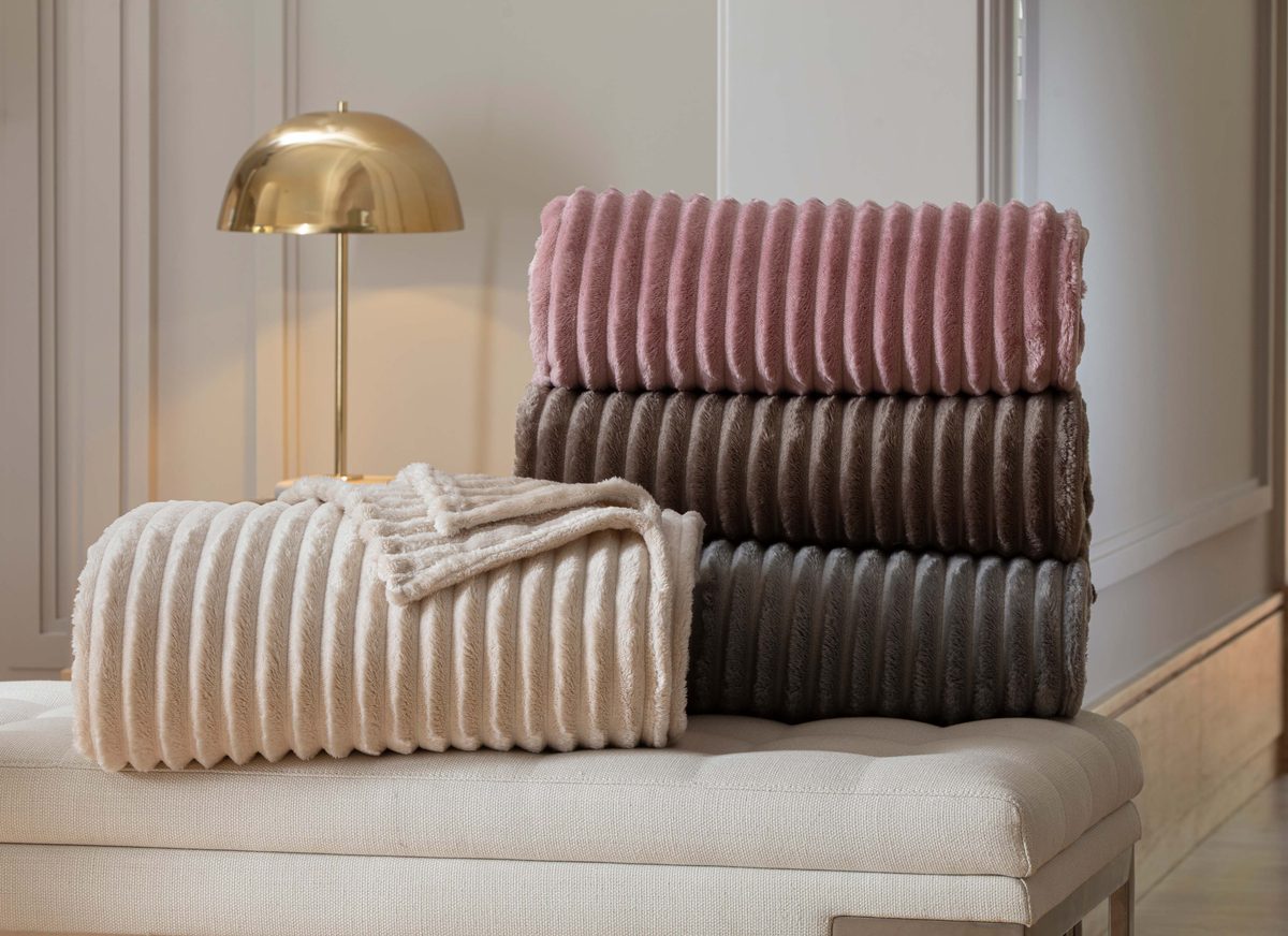 Pilha de cobertores Corttex London canelados nas cores rosé, marrom e cinza, e um cobertor bege dobrado sobre um banco