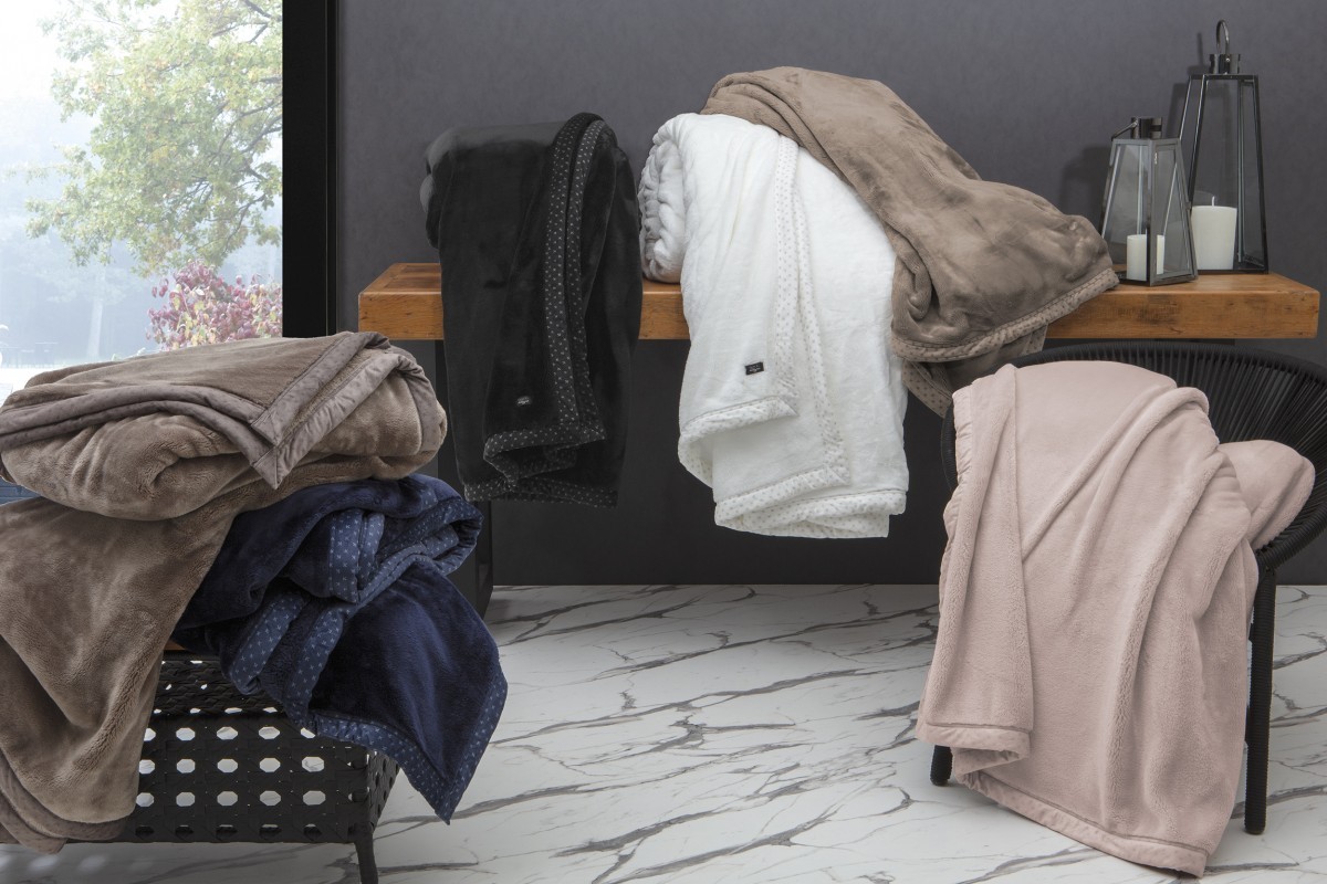Cobertores Kacyumara Hight 700 pendurados e dobrados sobre uma bancada, em preto, branco, bege e rosé, com borda estampada